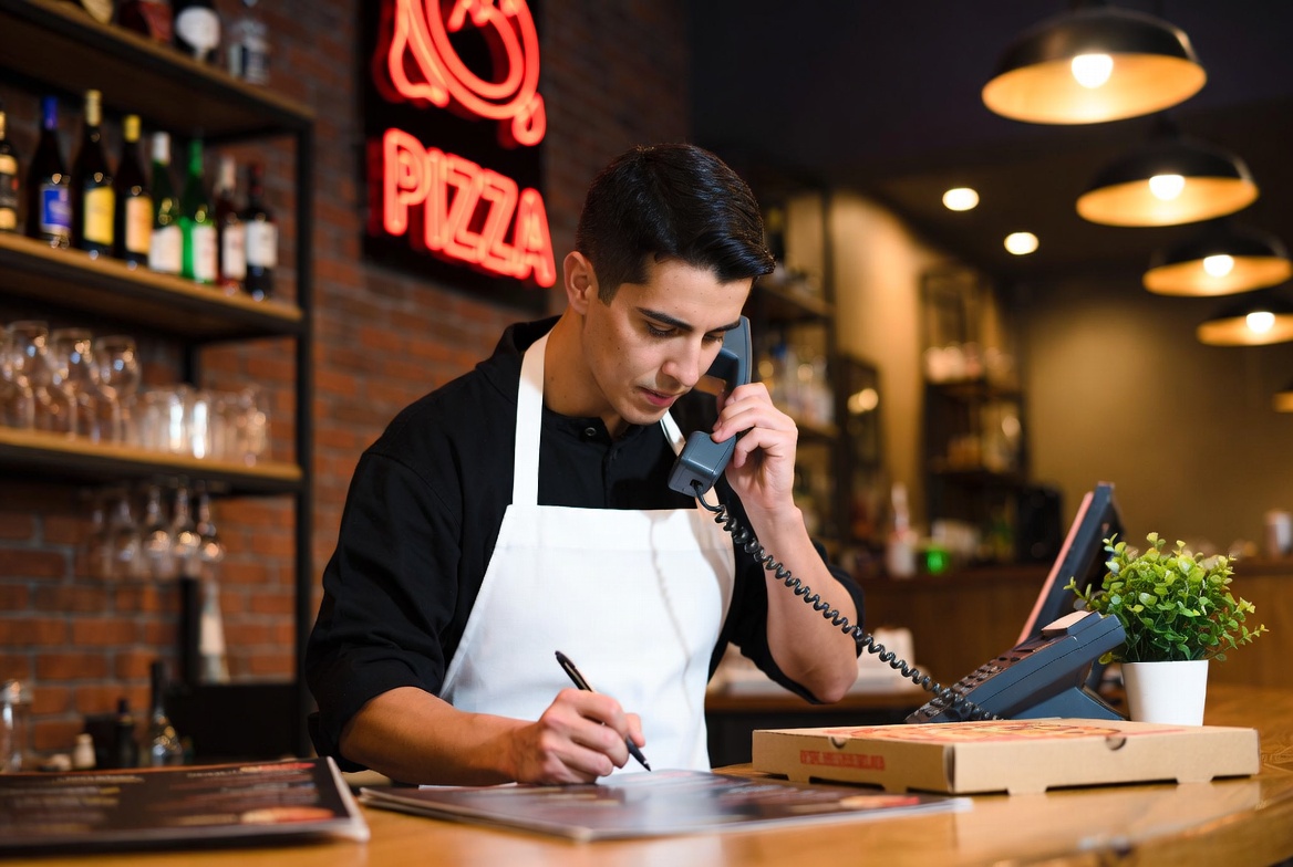 Restaurant worker taking a phone order at counter with pizza boxes and neon sign – representing how Level POS caller ID and saved cards speed up takeout orders